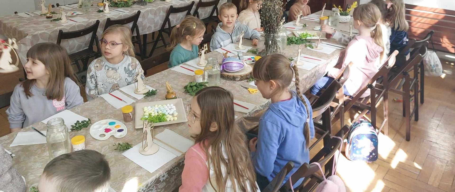 Dzieci siedzą przy długim stole, na którym są kartki i kartonowe zające, zielone gałązki