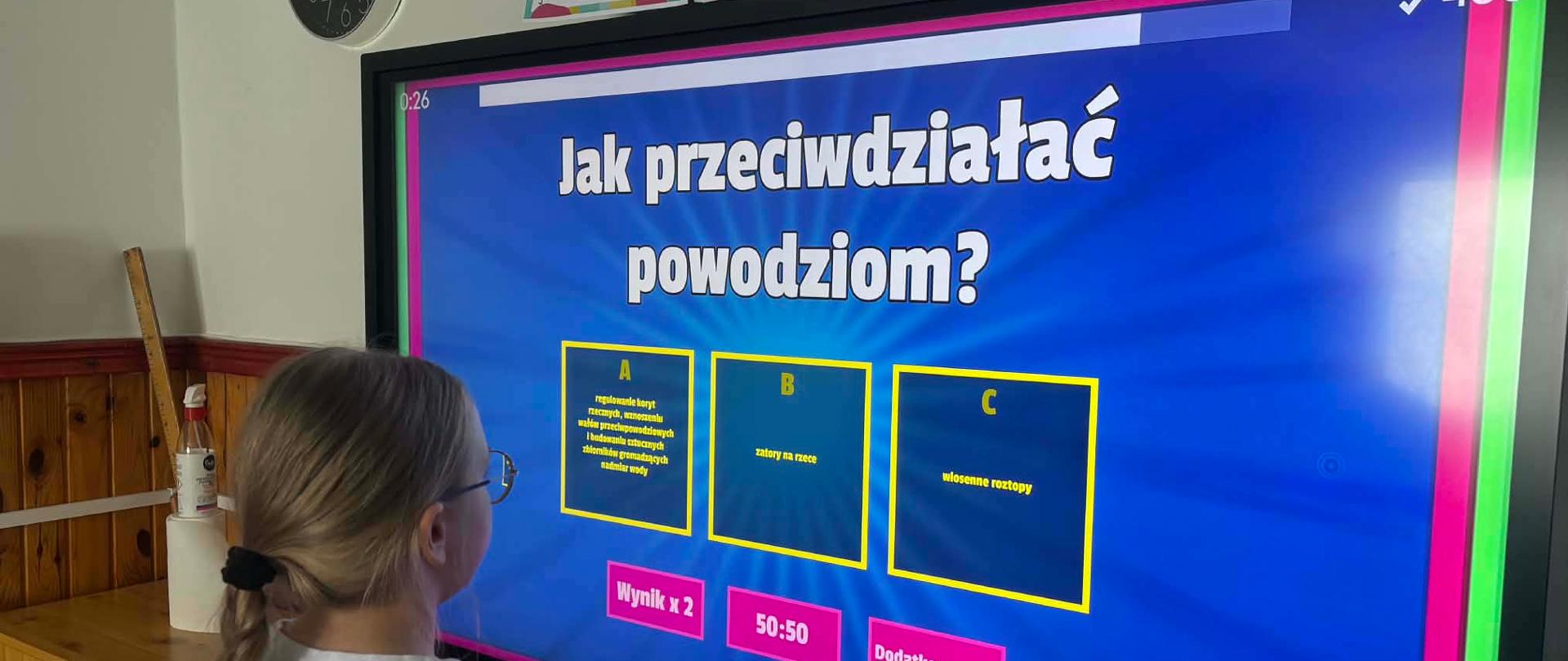 Uczennica klasy czwartej stoi przed monitorem interaktywnym. Na ekranie wyświetlony jest quiz z pytaniem: ,,Jak przeciwdziałać powodziom?". Pod spodem znajdują się trzy odpowiedzi. 