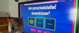 Uczennica klasy czwartej stoi przed monitorem interaktywnym. Na ekranie wyświetlony jest quiz z pytaniem: ,,Jak przeciwdziałać powodziom?". Pod spodem znajdują się trzy odpowiedzi. 