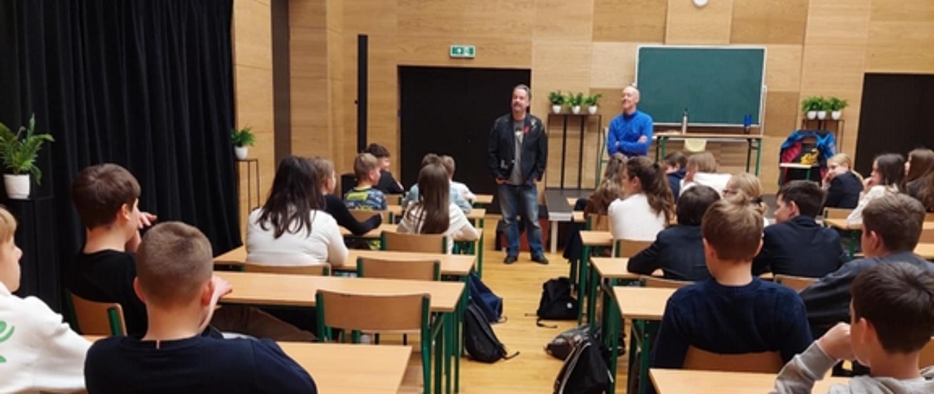 Grupa uczniów, widziana od tyłu, siedzi w ławkach w jednej z sal Teatru Ludowego. Przed nimi stoją reżyser oraz aktor mówiąc coś do zebranych młodych gości.