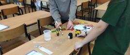 Dwie osoby stoją przy stoliku. Jedna z osób ma mazak w ręku a druga baterię