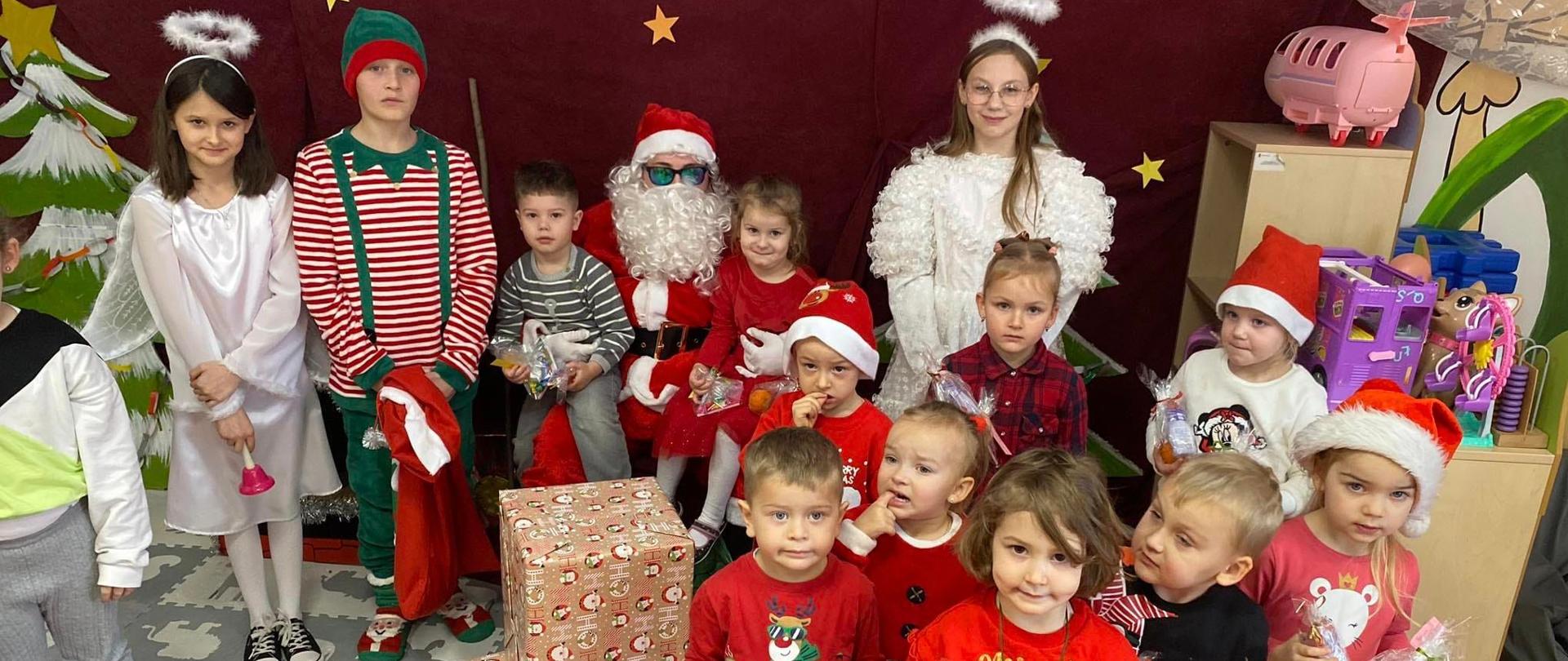 Przedszkolaki pozują do zdjęcia z prezentami otrzymanymi od Mikołaja, z tyłu siedzi Mikołaj, trzyma dziewczynkę i chłopca na kolanach, po bokach stoją Aniołki i Elf