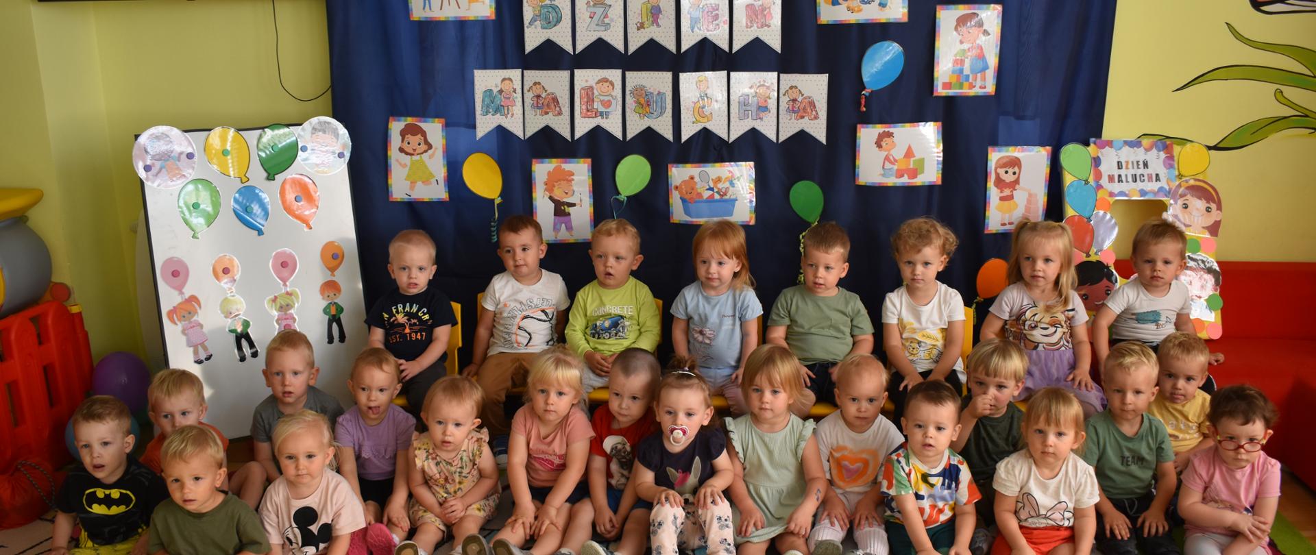 Na zdjęciu widać grupę 25 dzieci, siedzących w dwóch rzędach, a w tle jest niebieski materiał oraz dekoracje związane z dniem malucha.