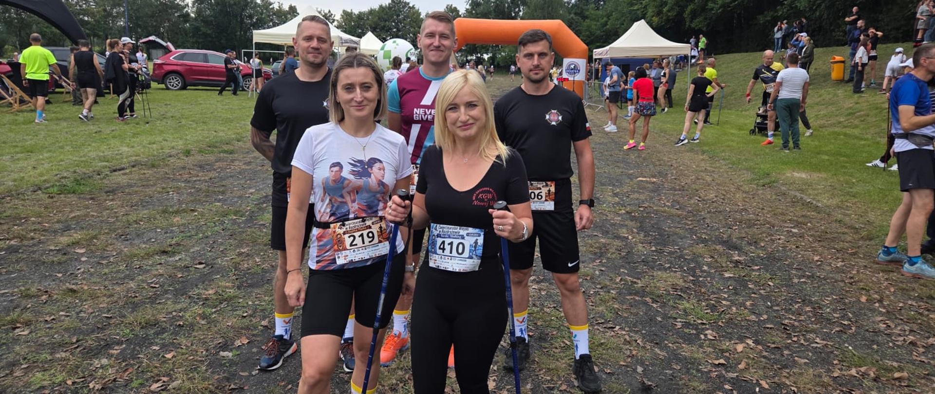 Dwie kobiety i trzech mężczyzn w strojach sportowych, na bluzkach widać przypięte numery. Kobieta po prawej stronie trzyma kijki do nordic walking. Za nimi samochody, namioty, tłum ludzi. W tle widać wysokie drzewa.