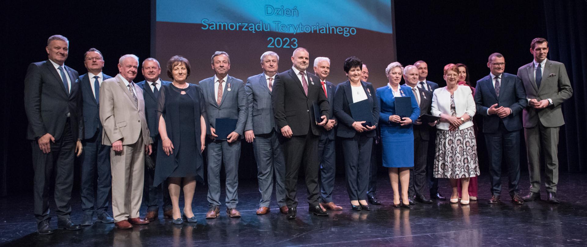 Zdjęcie grupowe na scenie Sokołowskiego Ośrodka Kultury z udziałem osób odznaczonych oraz parlamentarzystów. W tle na ekranie powiewa biało - czerwona flaga z napisem Dzień Samorządu Terytorialnego.
