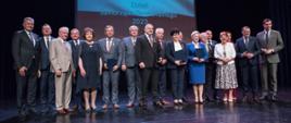 Zdjęcie grupowe na scenie Sokołowskiego Ośrodka Kultury z udziałem osób odznaczonych oraz parlamentarzystów. W tle na ekranie powiewa biało - czerwona flaga z napisem Dzień Samorządu Terytorialnego.