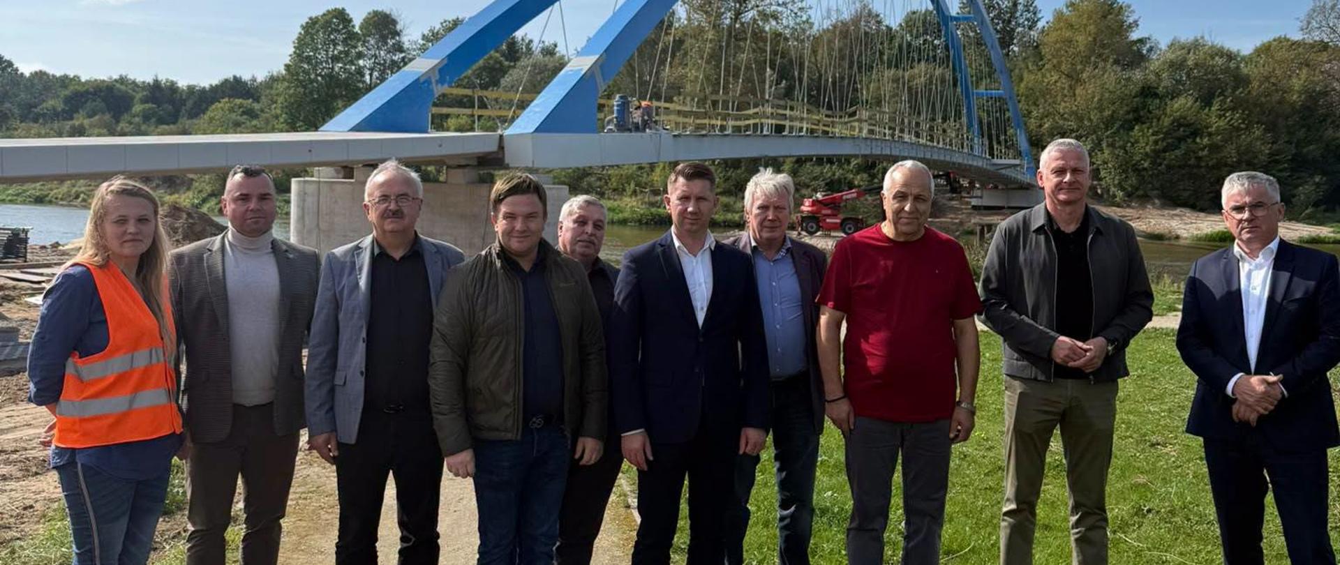 Grupa dzieisięciu osób pozuje nad rzeką, za którą znajduje się niebieski łukowy most i porośnięty drzewami krajobraz.
