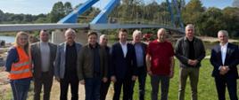 Grupa dzieisięciu osób pozuje nad rzeką, za którą znajduje się niebieski łukowy most i porośnięty drzewami krajobraz.