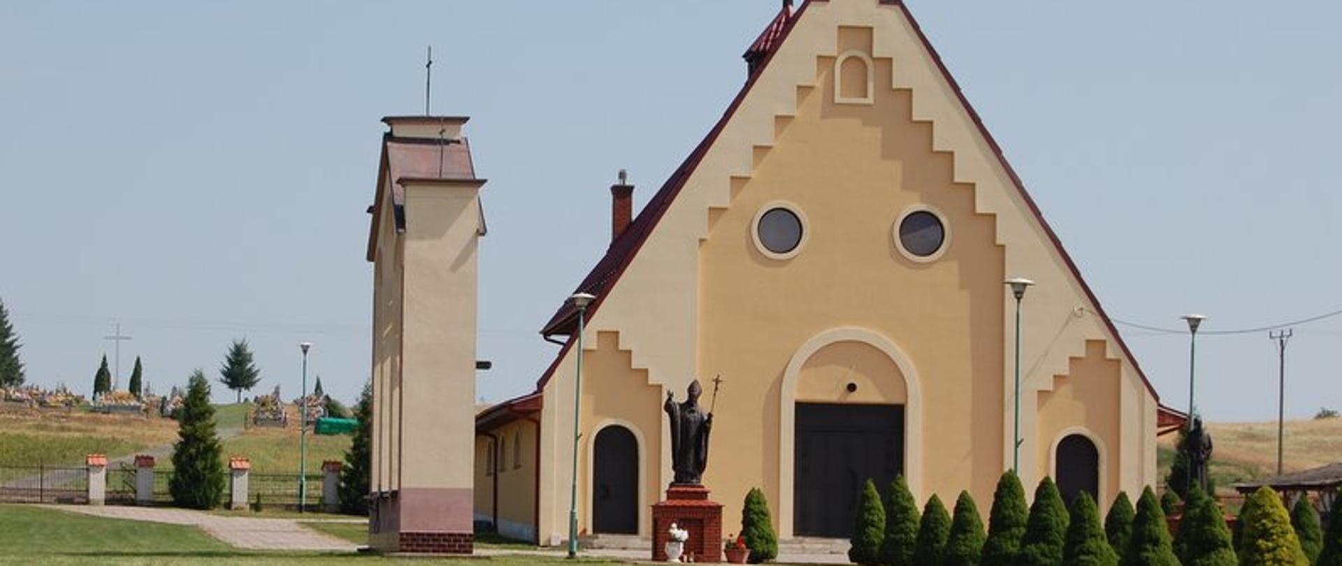 Bezowy kościół z czerwonym dachem i krzyżem na dachu. Przed kościołem figura Jana Pawła II i posadzone zielone drzewka. 