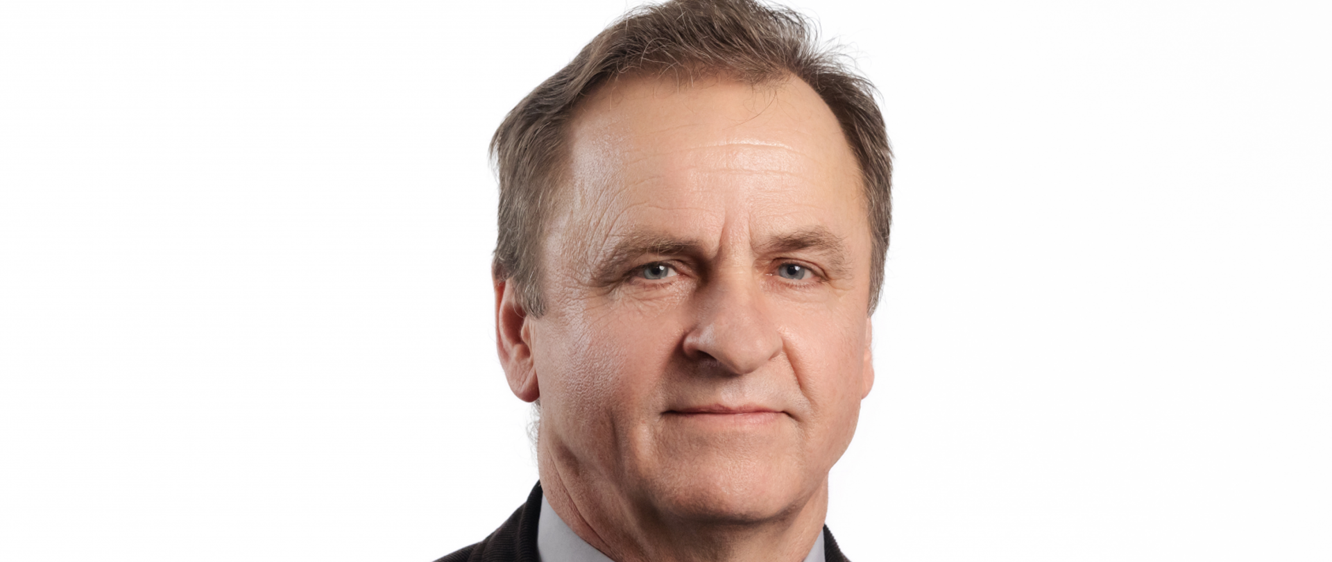 Starszy mężczyzna, szatyn, włosy zaczesane do tyłu, niebieskie oczy. Ubrany w ciemnobrązową marynarkę, szara koszulę i stalowy krawat