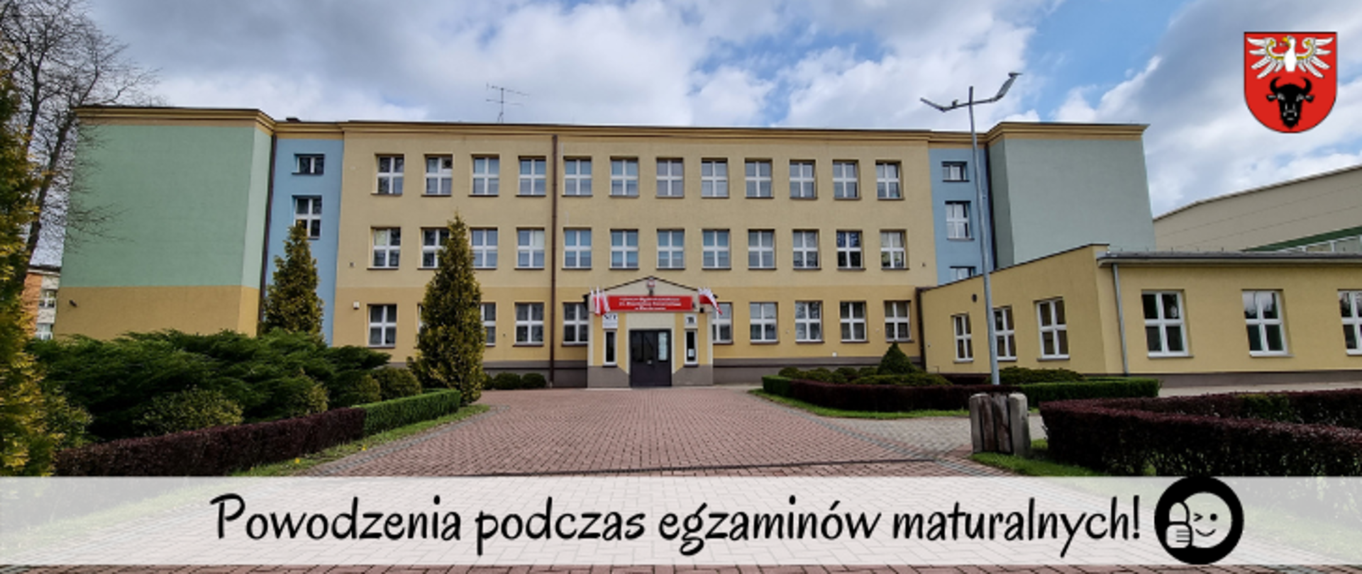 na dole znajduje się napis :"Powodzenia podczas egzaminów maturalnych!" obok napisu emotka z ksiukiem, w tle budynek ILO w Zambrowie, a w górnym prawym rogu logo powiatu zambrowskiego