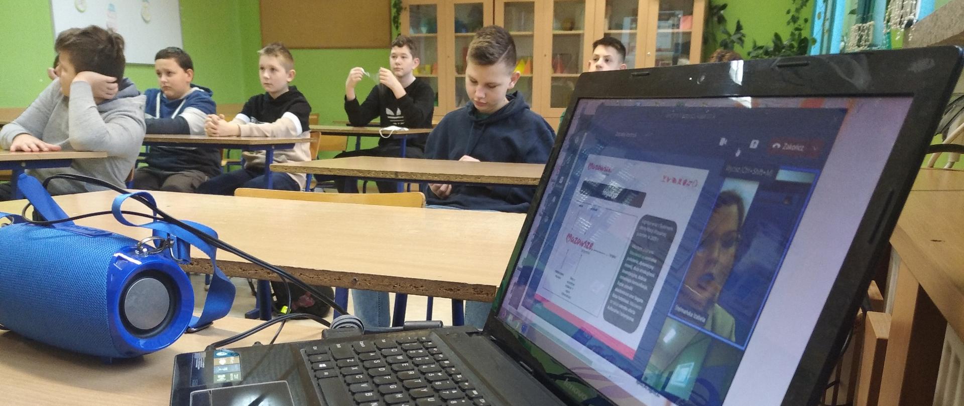 Młodzież siedzi przy ławkach szkolnych. Na pierwszym planie włączony laptop