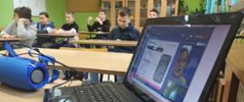 Młodzież siedzi przy ławkach szkolnych. Na pierwszym planie włączony laptop