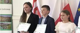 Dwie uczennice i jeden uczeń biorący udział w konkursie trzymają w ręku dyplomy, za nimi flaga gminy, Polski i Unii Europejskiej
