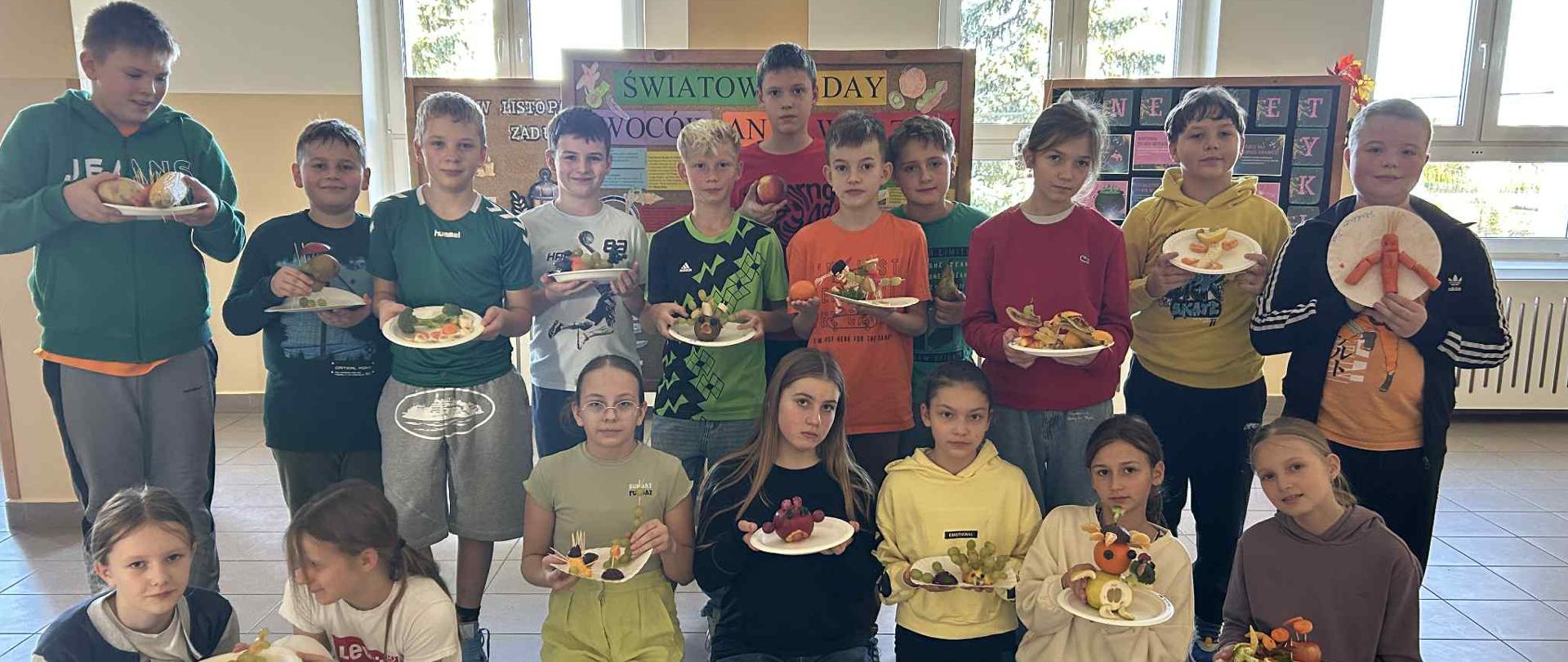Grupa uczniów ubranych w kolorowe ubrania trzymających w rękach przygotowane przez siebie talerze ,, owocowo- warzywne"