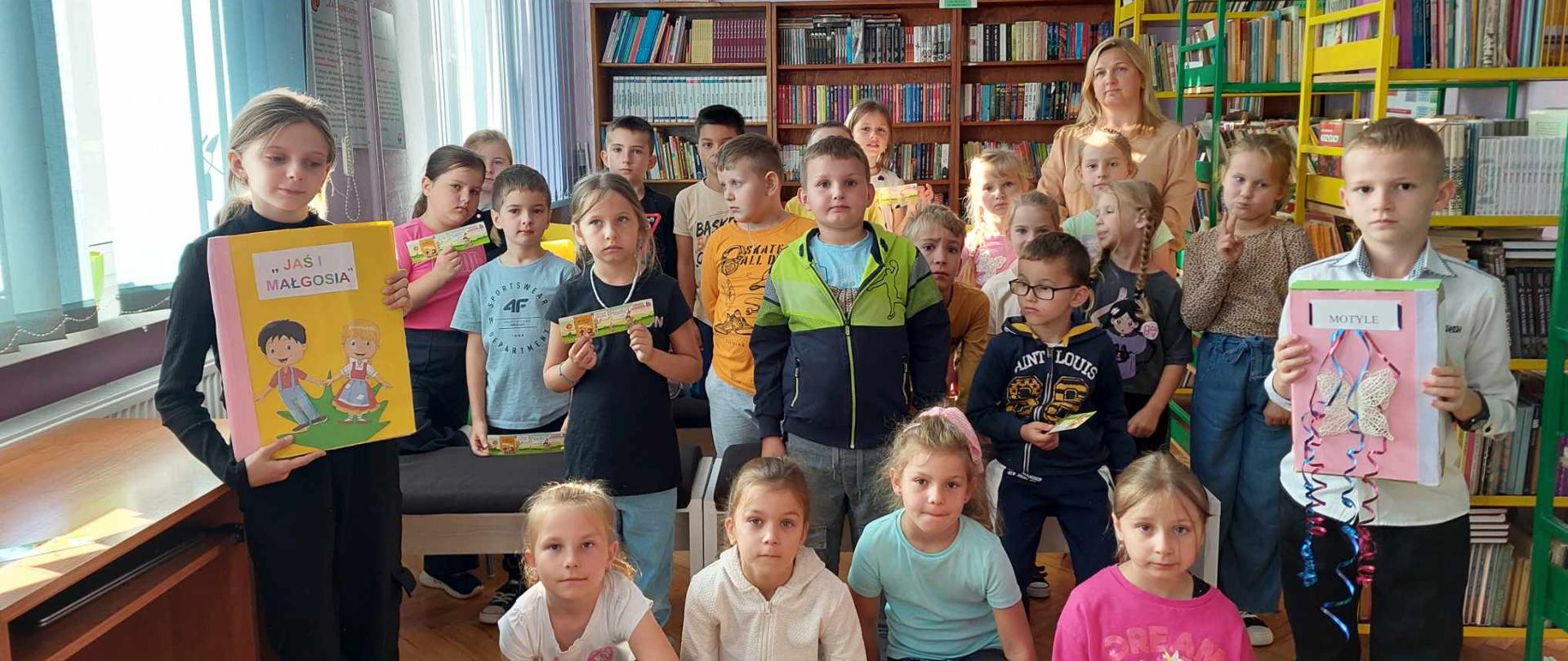 Uczniowie klasy pierwszej pozują do zdjęcia w szkolnej bibliotece. Po lewej stronie dziewczynka, a po prawej chłopiec z klasy piątej trzymający w rękach książki. W tyle pani bibliotekarka