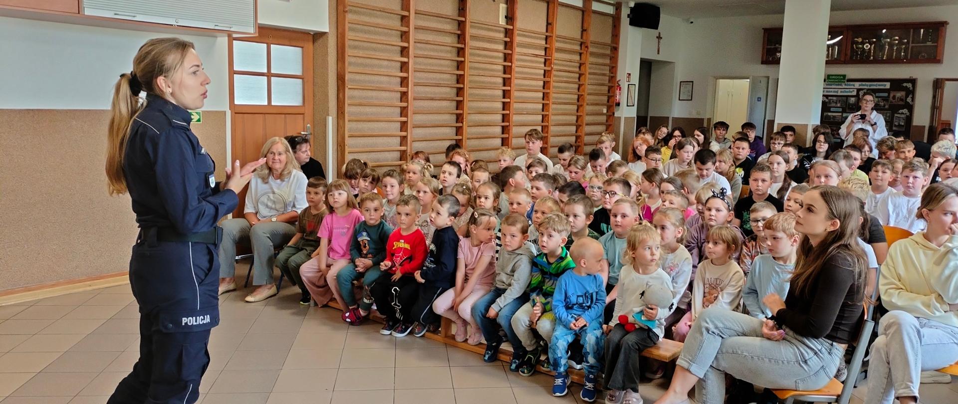 Pani policjant stoi na głównym korytarzu szkoły. Przed nią na ławeczkach oraz krzesłach siedzą uczniowie. Ich twarze skierowane są ku pani funkcjonariusz.