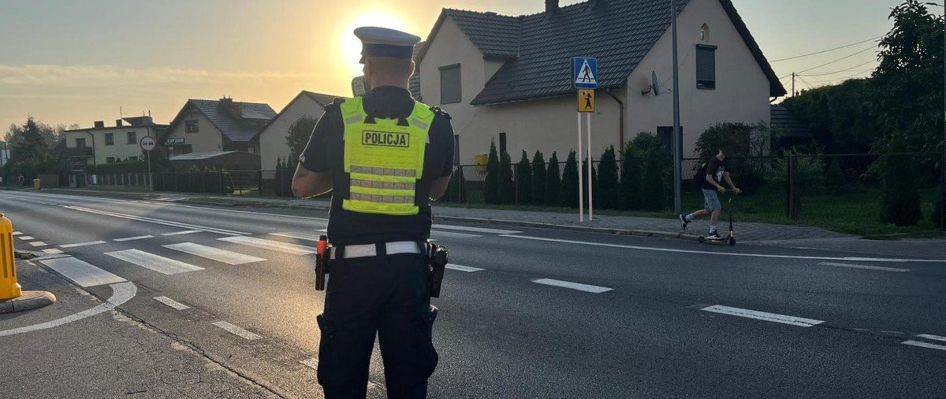 Policjant w żółtej kamizelce stojący przy drodze. W tle domki jednorodzinne.
