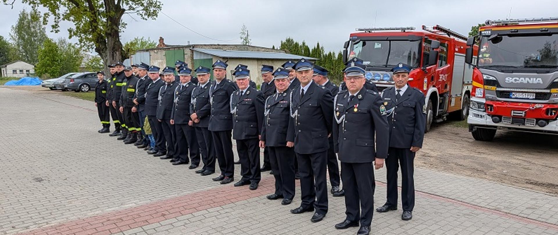grupa osób ubranych w mundury strażackie w dwuszeregu. Za nimi samochody strażackie- bojowe.