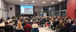 Szerokie ujęcie sali konferencyjnej, z większością uczestników siedzących tyłem do obiektywu. Na ekranie w głębi wyświetlana jest mapa przebiegu drogi ekspresowej S7. Kobieta stoi i przemawia pod ekranem. Po prawej stronie widać stojak z napisem "GMINNY PUNKT" i ozdobną choinkę.
