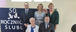 Państwo Krystyna i Zenon na tle granatowej tablicy z napisem wraz z Wójtem, Z-cą Wójta i Z-cą Kierownika USC