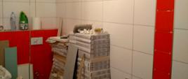 pomieszczenie toalety, na ścianach płytki w kolorach białym i czerwonym, przy ścianie stoją materiały i narzędzia budowlane