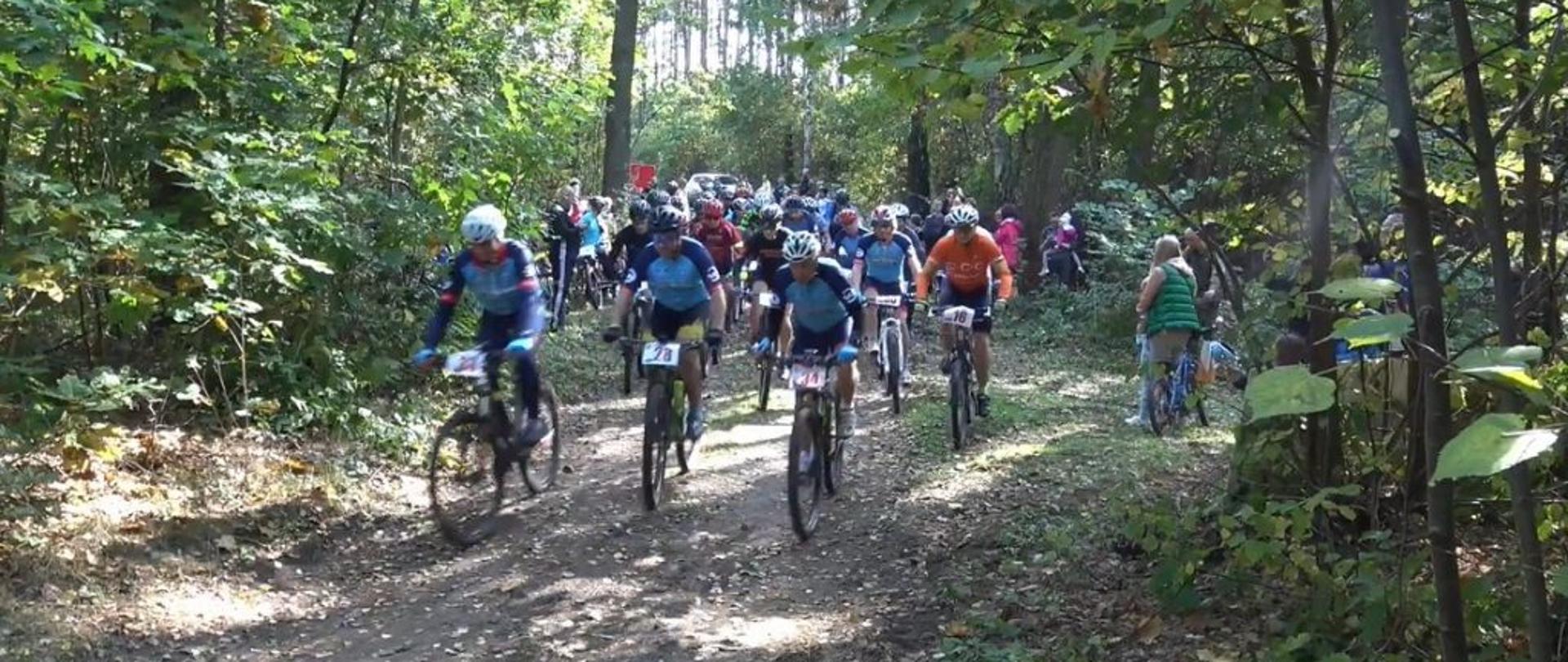 Uczestnicy rajdu w trakcie rywalizacji – kilkudziesięciu uczestników na rowerach, w strojach sportowych. W tle las, w którym odbywają się zawody.