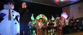 Dyrygent oraz grupa dzieci z instrumentami podczas występu na scenie. W tle widać dekoracje świąteczne.