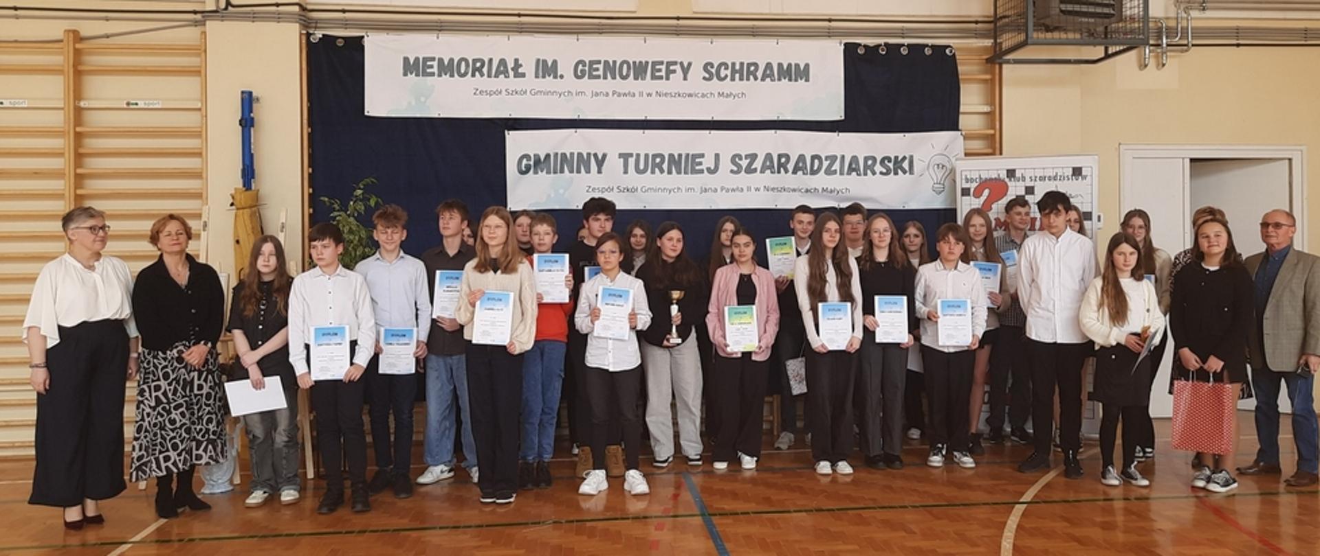 W szkolnej sali gimnastycznej do zdjęcia pozuje grupa uczniów trzymająca w dłoniach Dyplomy. Po lewej stronie grupy stoją dwie kobiety, a po prawej jeden mężczyzna.