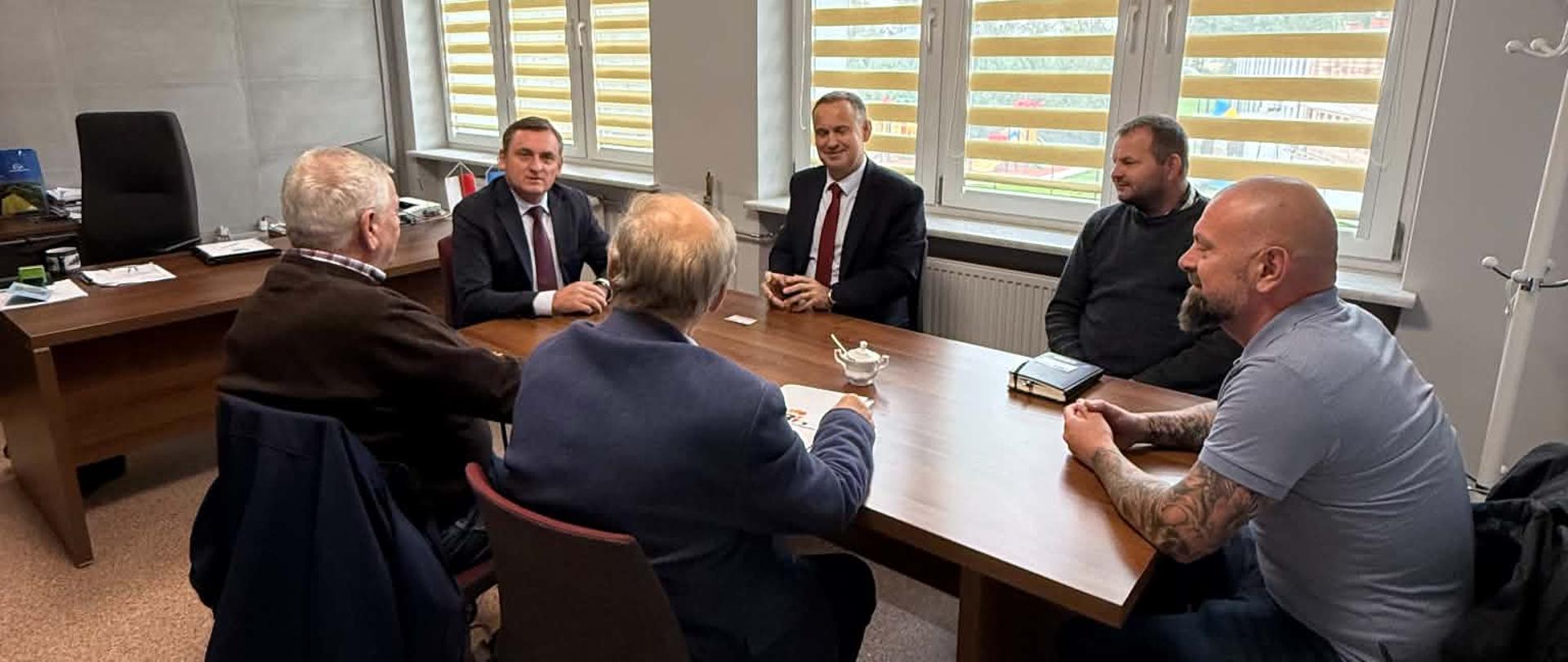 Uczestnicy narady siedzą wokół stołu w biurze; na ścianie w tle widoczne jest godło Polski.