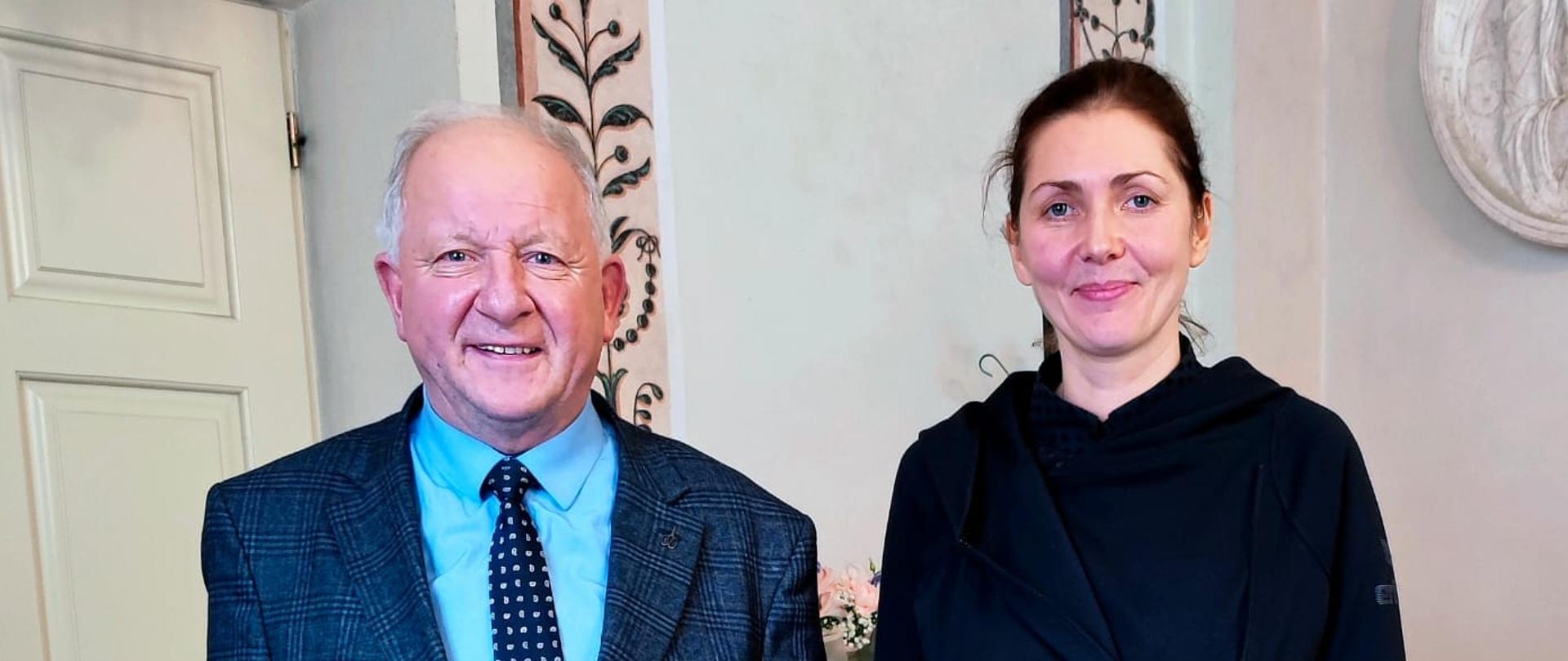 W biurze stoją dwie osoby po lewej mężczyzna w garniturze niebieskiej koszuli i krawacie w kropki po prawej kobieta z czarnymi włosami, czarnej bluzie i granatowych spodniach.