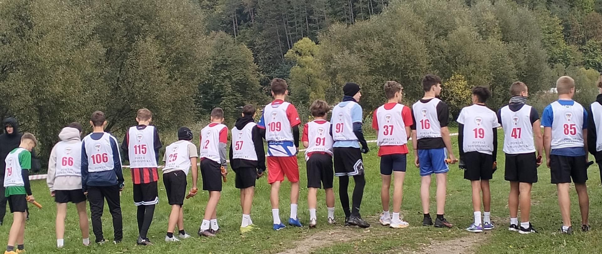 Na trawiastej trasie biegowej stoją chłopcy w białych koszulkach z numerami nałożonych na własne stroje sportowe, przygotowani do biegu, pokazani są od tyłu, w tle drzewa i krzewy