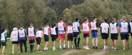 Na trawiastej trasie biegowej stoją chłopcy w białych koszulkach z numerami nałożonych na własne stroje sportowe, przygotowani do biegu, pokazani są od tyłu, w tle drzewa i krzewy
