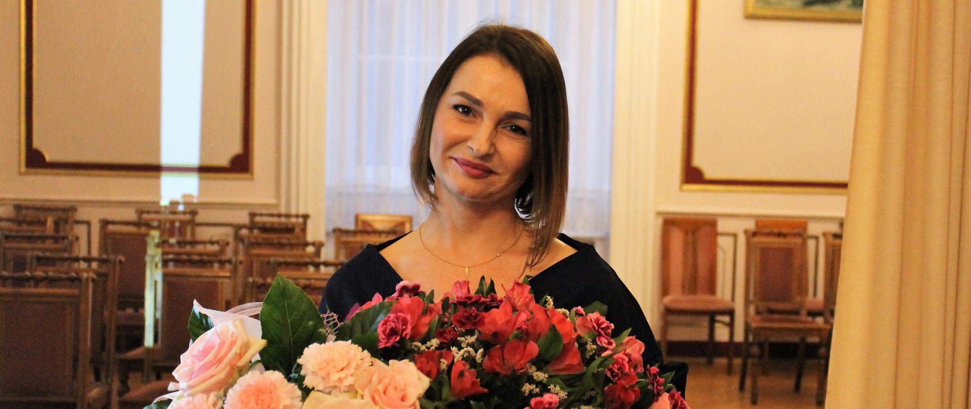 Ewa Bączkowska - Skarbnik Gminy Świerklaniec - młoda kobieta o ciemnych włosach do ramion, uśmiechnięta z dwoma bukietami kwiatów w dłoniach
