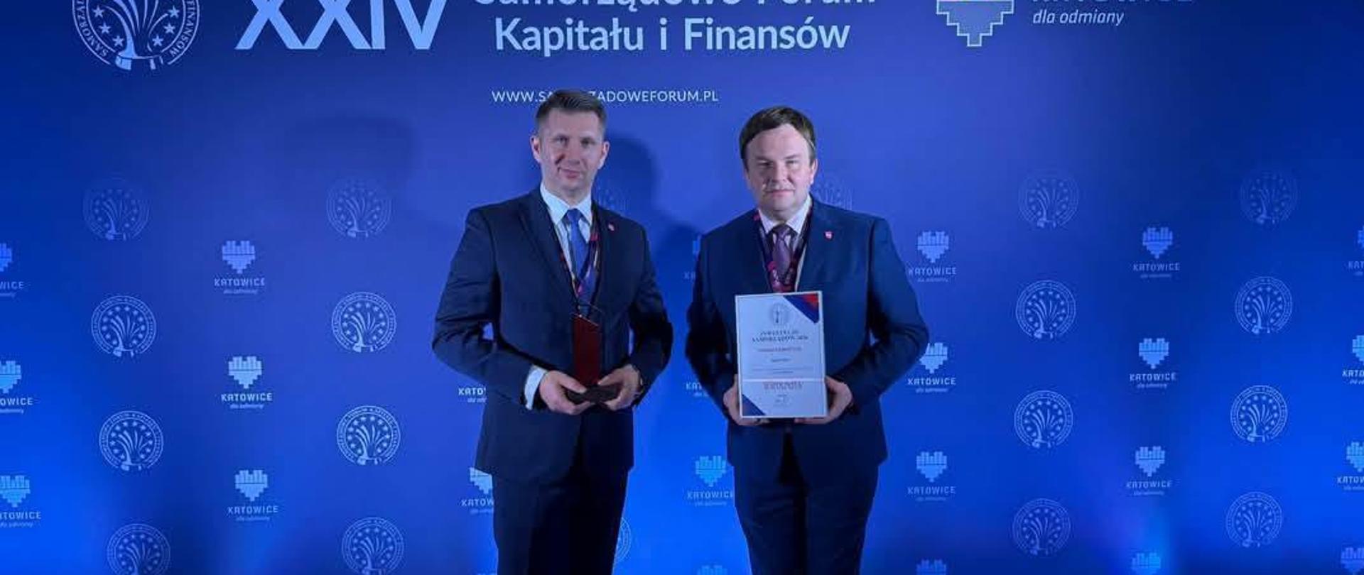 Dwóch mężczyzn w strojach wieczorowych trzymających nagrody na scenie z niebieskim tłem z logo.