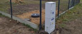 Obraz przedstawia ogrodzony teren, na którym znajduje się biały rozdzielnik elektryczny umieszczony na zewnątrz ogrodzenia. Ogrodzenie wykonane jest z metalowej siatki i ma betonową podstawę. Wewnątrz ogrodzonego obszaru widoczny jest okrągły, czarny obiekt leżący na ziemi, pokrywa studzienki oraz kopiec ziemi.