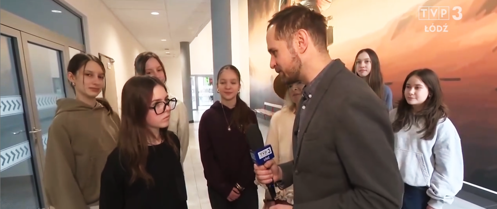 Reporter TVP3 Łódź przeprowadza wywiad z grupą nastolatek w holu budynku. Dziennikarz trzyma mikrofon z logo TVP3, a młodzież stoi półkolem i odpowiada na pytania.