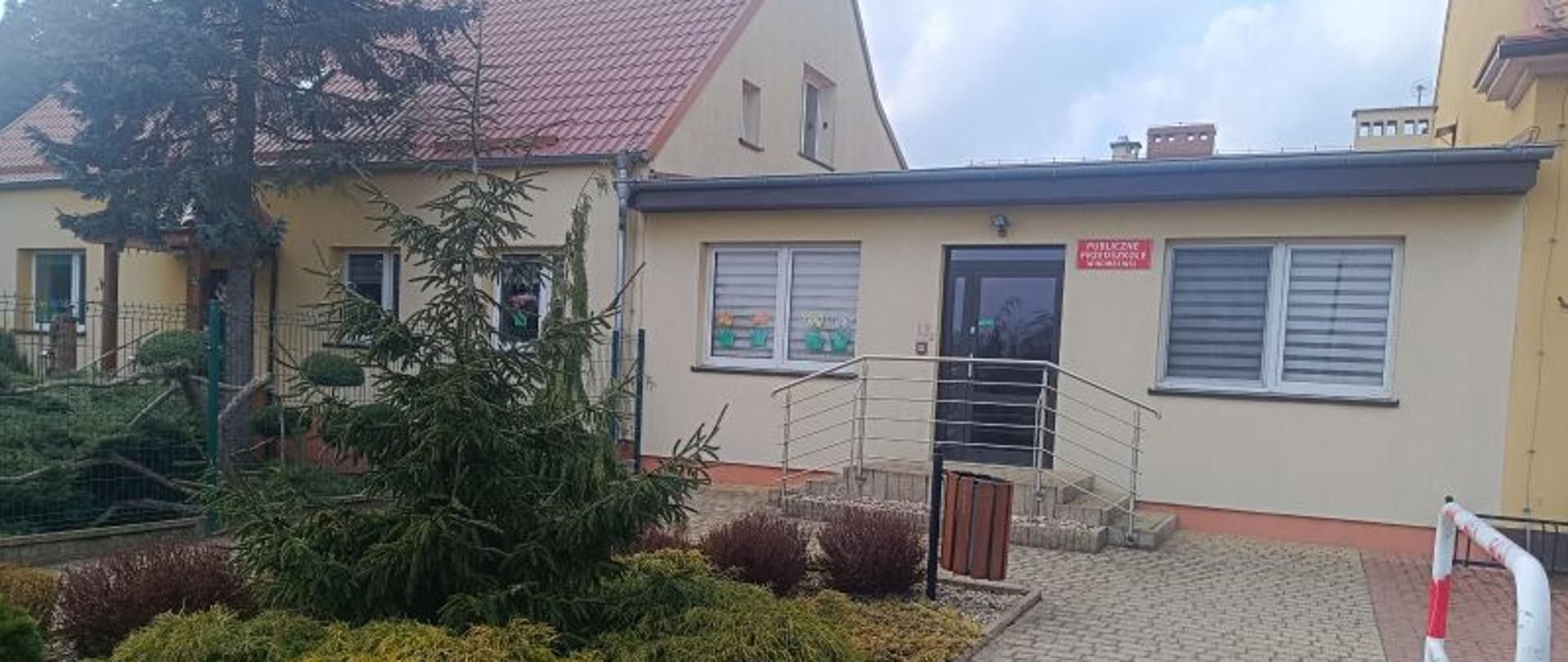 Dwa budynki z wysokimi dachami pokrytymi czerwoną blachą, między nimi niski łącznik a w nim wejście do budynku a po prawej i lewej stronie okna. Do budynku prowadzą schody ze stopniami po prawej i lewej stronie. Po prawej stronie drzwi tabliczka Publiczne Przedszkole w Nowej Wsi. Przed budynkiem zieleń, po prawej stronie barierka. 