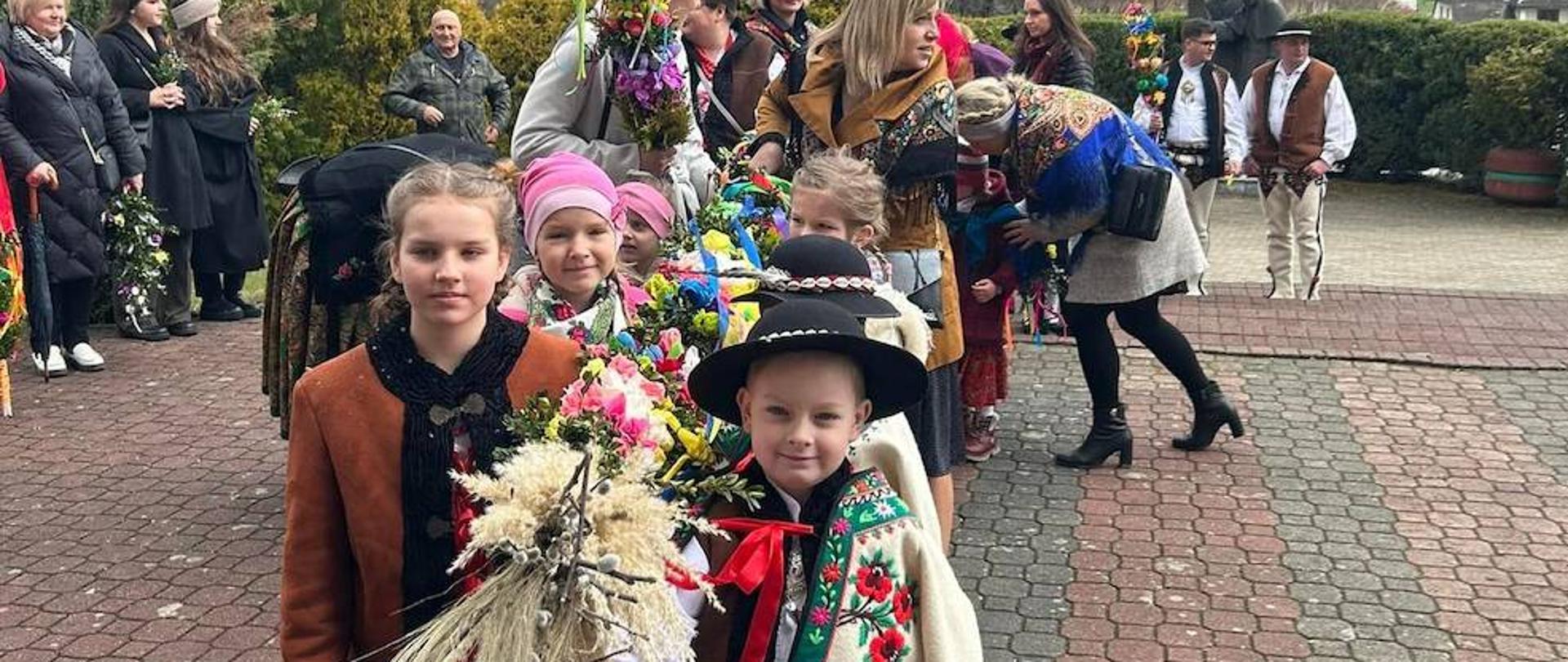 Zdjęcie przedstawia dzieci ubrane w stroje regionalne trzymające jedną, dużą palmę wielkanocną