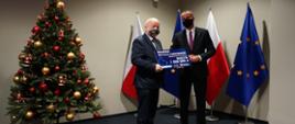 Wojewoda i Burmistrz trzymający symboliczny czek, w tle flagi Polski oraz Unii Europejskiej, z lewej strony udekorowana choinka