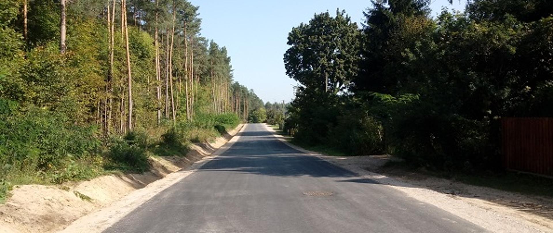 Na zdjęciu widoczna nowo wybudowana droga asfaltowa w Trzciance. Po lewej stronie drogi znajduje się rów oraz las. Po lewej stronie widać płot ogradzający posesję oraz drzewa.
