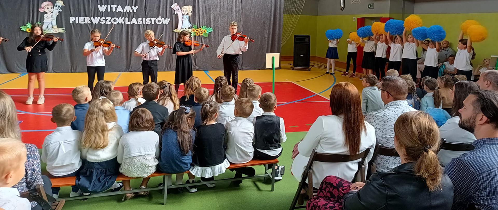 Uczniowie grający na skrzypcach oglądani przez dzieci szkolne i rodziców.