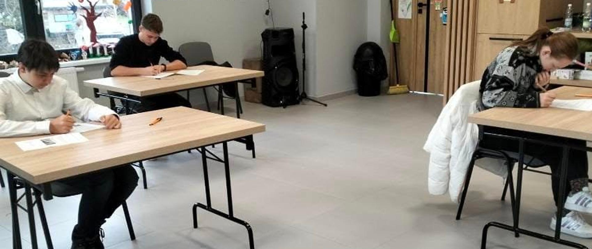 Jedna uczennica i dwóch uczniów siedzą pojedynczo przy stolikach i piszą arkusz konkursowy, przestronna jasna sala, na ścianie prace plastyczne, drewniane szafki