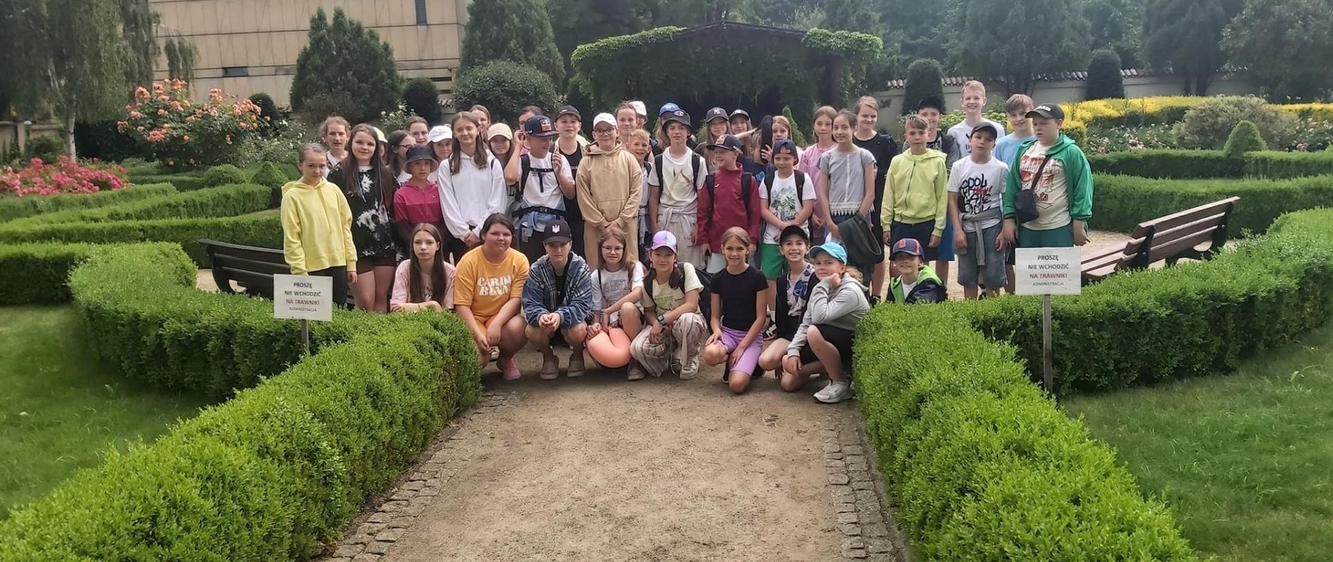 Uczniowie klas IV- VIII wspólnie ustawieni na tle W=wrocławskiego ogrodu. W tle dużo zieleni, na pierwszym planie widoczna ścieżka dla zwiedzających ogrody.