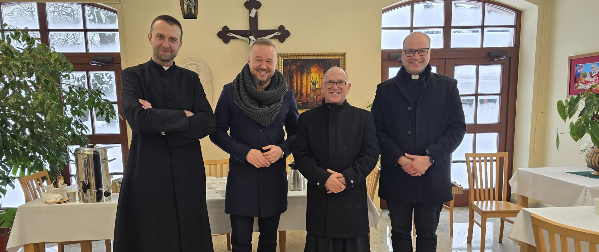 Mężczyźni pozują do zdjęcia. W tle żółta ściana na której wisi krzyż