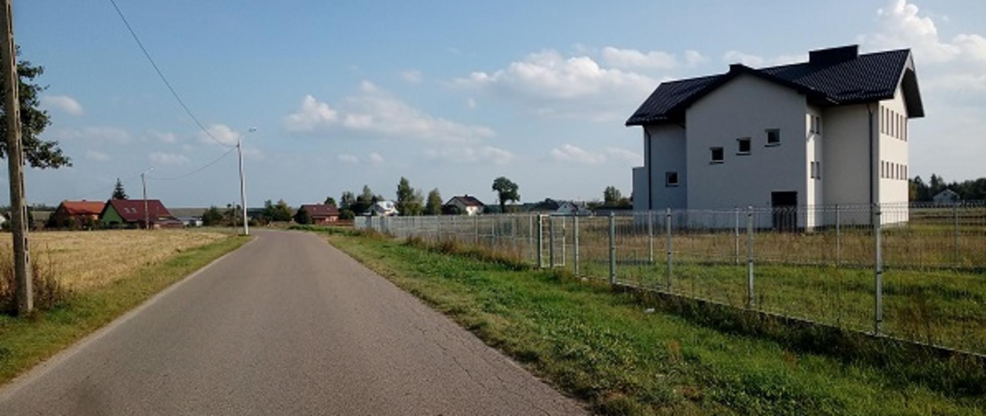 Na zdjęciu po prawej stronie widoczny biały budynek OSP Białebłoto, działki ogrodzone panelami stalowymi. W lewej części zdjęcia widoczna jest wiejska droga.