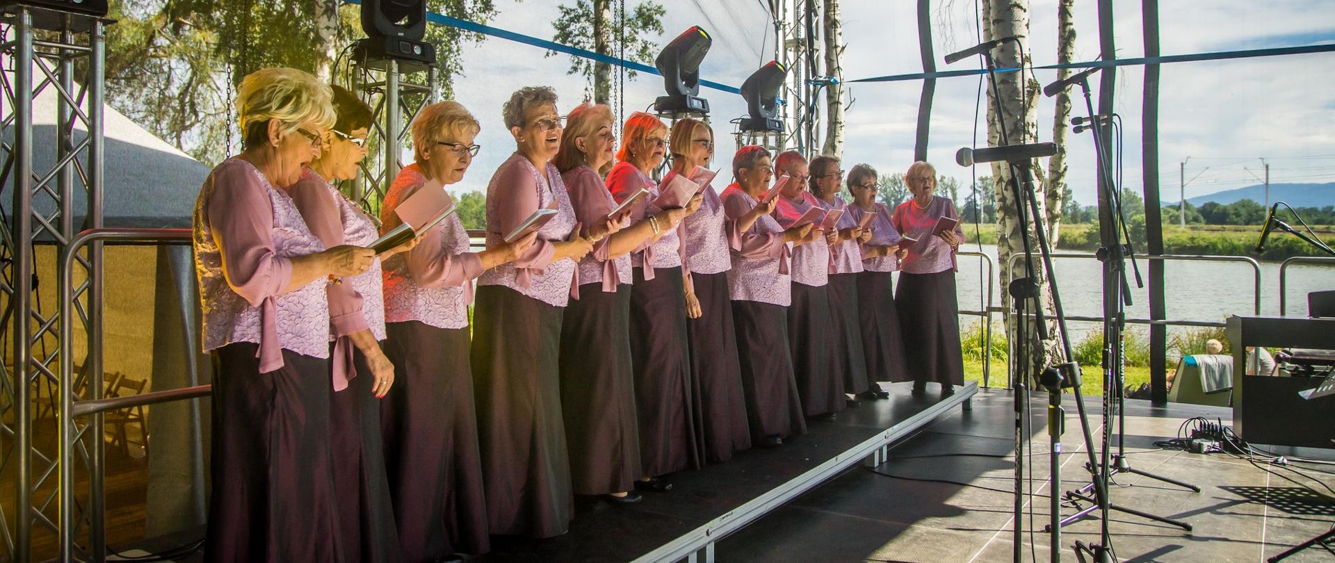Kobiety w różowo brązowych strojach śpiewają na scenie