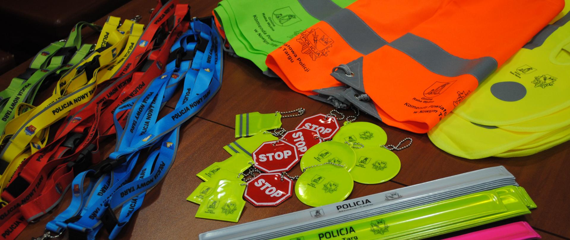 Na stole leżą gadżety odblaskowe takie jak: opaski, breloczki, kamizelki i worki. 
