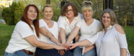 Kobiety w białych koszulach i bluzkach wyciągają rękę do środka na znak
przymierza. Jest ich pięć, są uśmiechnięte.