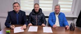 Trzech mężczyzn siedzi przy drewnianym stole w biurze; przed nimi leżą dokumenty, a jeden z nich je podpisuje